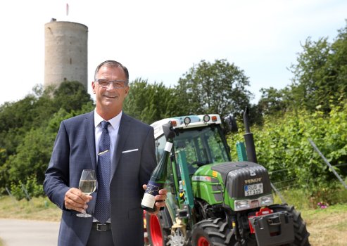 Bürgermeister Winfried Steinmacher mit blauer Brille steht mit blauem Anzug mit weißem Einstecktuch, dunklem Gürtel, weißem Hemd, blauer Krawatte und silberner Krawattennadel mit gefülltem Weinglas und Weinflasche in der Hand vor einem Traktor neben dem Weinberg der Ehe. Im Hintergrund befindet sich die Burgruine Scharfenstein mit weiß-roter Fahne, Laubbäumen und ein weißer Himmel. 