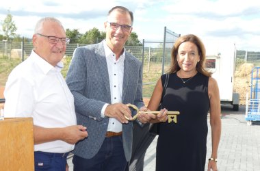 links Rüdger Wolf in blauer Hose und weißem Hemd mit Brille, in der MItte Winfried Steinmacher mit blauer Hose, braunem Gürtel, weißem Hemd mit schwarzen Knöpfen und blau weiß kariertem Sakko und Brille. Rechts Brigitte Pietsch in einem schwarzem Kleid ohne Arm. Im Hintergrund Gitter-Zäune aus Metall und ein blauer Himmel mit Wolken.   