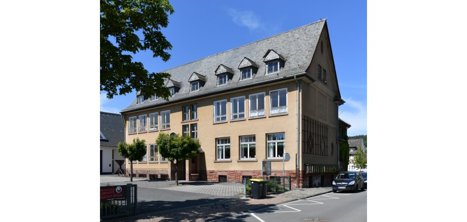 Beiges Gebäude mit Fenstern und Dachfenstern in dem mit schwarzen Ziegeln bedeckten Dach, In dem Gebäude befand sich früher die Schule von Kiedrich. Ebenso sieht man den Hof mit Bäumen und ein Teil der Straße mit einem parkendem Auto. 