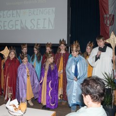 Die Kiedricher Sternsinger, elf Kinder, mit Kronen auf dem Kopf und langen farbigen Umhängen, sowie ein Messdiender mit weiß-schwarzem Gewand, einem langem Stock mit Stern und einem Mikrofon in der Hand, stehen vor einer Leinwand mit dem Text "Segen bringen, Segen sein" und rechts davon eine Fahne in rot-weiß, mit dem Kiedricher Wappen.