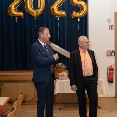 Bürgermeister Steinmacher (links) mit dem Ersten Beigeordneten Rüdiger Wolf (rechts), stehen, beide im dunklen Anzug und mit Brille, vor goldenen Zahlenballons die 2025 zeigen, vor einem blauem Vorhang.