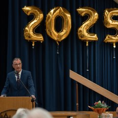 Bürgermeister Steinmacher, in blauem Anzug, mit weißem Hemd, Kravatte und blauer Brille  steht hinter einem Mikrofon an einem Rednerpult. Im Hintergrund hängt die rot-weiße Fahne mit Kiedricher Wappen und goldenen Zahlenballons die 2025 zeigen, vor einem blauem Vorhang.