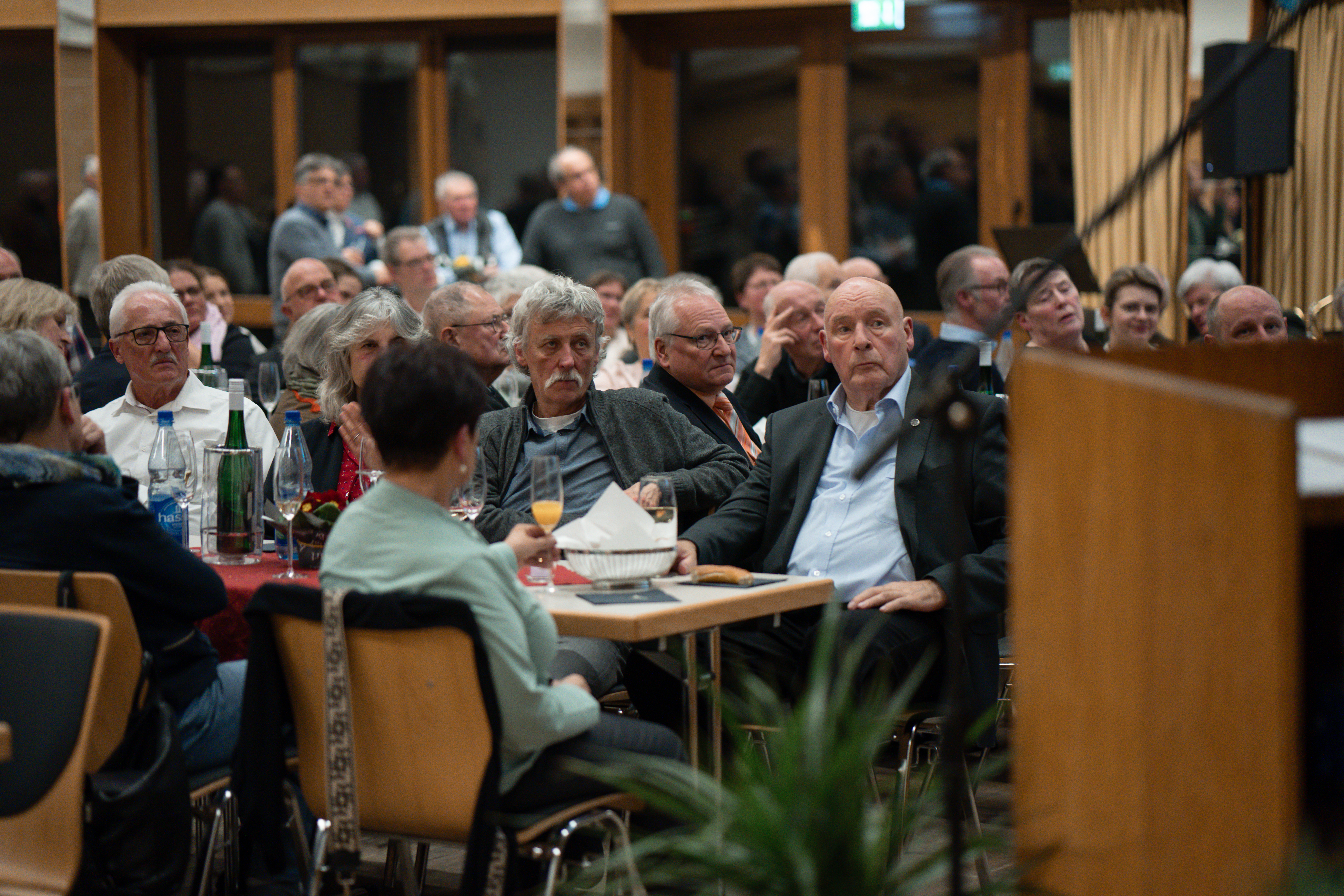 Man sieht das Publikum des Neujahrsempfangs an Tischen, auf Stühlen sitzen. Im Hintergrund steht das Publikum an Bistrotischen vor einer Fensterfront.