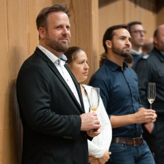 Vor einer Holzwand steht  Jochen Klein, Leiter der Kita Hickelhäusje und SPD-Vorsitzender im dunklem Anzug mit weißem Hemd. Neben dran, eine Frau in weiß gekleidet und im Hintergrund vier Männer, ebenso dunkel gekleidet.   