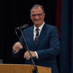 Bürgermeister Steinmacher, in blauem Anzug, mit weißem Hemd, Kravatte und blauer Brille  steht hinter einem Mikrofon an einem Rednerpult und lacht. Im Hintergrund ist ein blauer Vorhang.