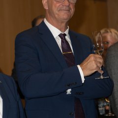 Bürgermeister Steinmacher, in blauem Anzug, mit weißem Hemd, Kravatte, blauer Brille und einem Sektglas in der Hand.