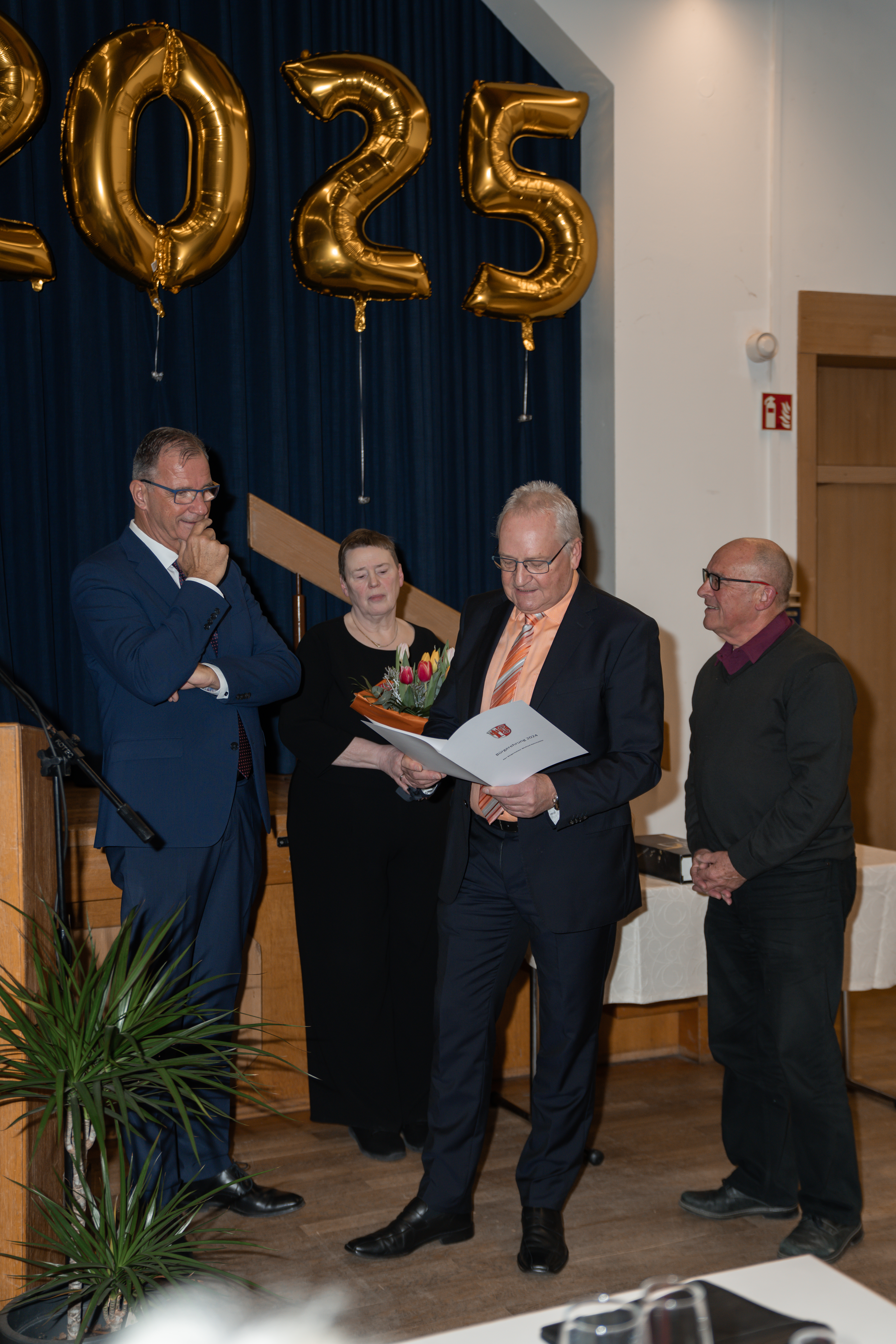 Bürgermeister Winfried Steinmacher (links), in blauem Anzug und weißem Hemd, mit Beate Schmidt, Vorsitzende der Gemeindevertretung, in schwarz gekleidet und einem Blumenstrauß mit Tulpen in der Hand, mit dem Ersten Beigeordneten Rüdiger Wolf, mit schwarzem Anzug, orangenem Hemd und gestreifter Kravatte und einer weißen Urkundenhülle mit dem Kiedricher Logo, in der Hand und der 2. Vorsitzendem des KCV Sprudelfunken Klaus-Peter Wiffler in schwarz gekleidet. Im HIntergrund sind goldenen Zahlenballons, die eine 2025 zeigen, vor einem blauem Vorhang.