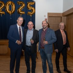 Bürgermeister Steinmacher (links), mit Walter Ruhl (Mitte) und dem Wallufer Bürgermeister Nikolaos Stavridis (rechts) und der Erste Beigeordneter Rüdiger Wolf (rechtaußen), alle im dunklen Anzug, stehen vor goldenen Zahlenballons, die 2025 zeigen, vor einem blauem Vorhang.