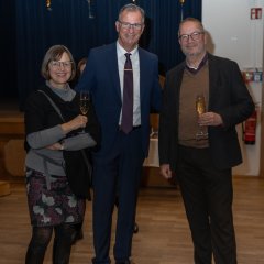 Bürgermeister Steinmacher (Mitte), mit dem ehemaligen Vorsitzenden der Gemeindevertretung Harald Rubel  mit seiner Ehefrau (links), alle im dunklen Anzug, die Damen trägt einen schwarzen MInirock mit grauen und weinriten Blumen und ein grauen Pullover mit einem schwarzen großen Schal. Alle tragen eine Brille und stehen vor goldenen Zahlenballons, die 2025 zeigen, vor einem blauem Vorhang.