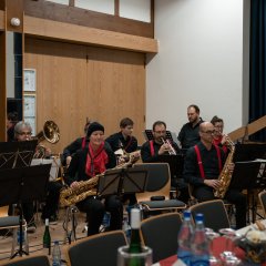 Die KCV Big Band, bestehend aus Männern und Frauen, in schwarz gekleidet, die Männer mit roten Hosenträgern und die Frauen mit roten Schals, sitzen auf Stühlen hinter Notenständern mit Liederblättern. In den Händen halten Sie Blasinstrumente, E-Gitarren. 