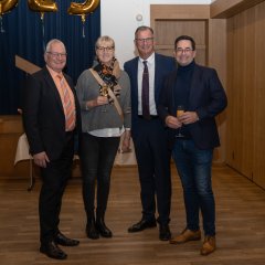 Erster Beigeordneter Rüdiger Wolf (links), Andreas Zeiselmaier, Vorstandsvorsitzender der Rheingauer Volksbank (rechts) mit seiner Ehefrau und Bürgermeister Winfried Steinmacher (Mitte), die Herren im dunklen Anzug und die Dame im grauen Pullover und einer schwarzen Hose, alle mit Brille, stehen vor einem blauem Vorhang und einer Holztür.