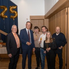 Erster Beigeordneter Rüdiger Wolf (links) und Bürgermeister Steinmacher (rechts davon, beide im dunklem Anzug mit Brille, daneben Fabian Sohlbach in beigem Hemd und dunkler Hose, Stefan Bibo in weißem Hemd und Jeans, Weinprinzessin Nikola Bibo, in schwarz gekleidetund mit Diadem in den blonden, langen Haaren und Georg Sohlbach (rechts), dunkel gekleidet, stehen alle vor goldenen Zahlenballons die 25 zeigen, vor einem blauem Vorhang und vor einer Holztür und einer Holzwand.