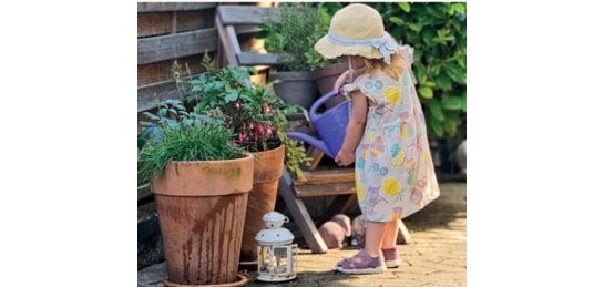 Ein kleines Mädchen im Blumenkleid und beigem Sonnenhut gießt mit einer blauen Gießkann zwei terrakottafarbene große Blumentöpfe, die mit Blumen bepflanzt sind, Davor steht eine weiße Laterne und im Hintergrund ein Holzstuhl, auf dem Blumentöpfe stehen und im Hintergrund eine belaubte Hecke.   