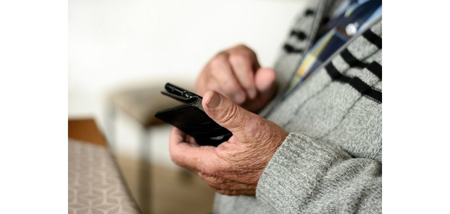 Man sieht den Oberkörper eine älteren Mannes, der ein blau-weiß-gelb karriertes Hemd trägt und eine grau-weiß-melierte Strickjacke mit schwarzen Steifen. In der Hand hält er ein Smart-Phone. Im HIntergrund steht ein Hocker und vor ihm ein Holzstisch mit einer beige-weiß gemusterten Tischdecke.