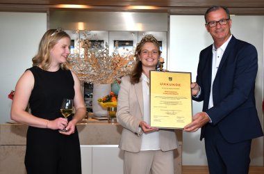 Die Kiedricher Weinprinzessin im schwarzen Kleid mit einem gefülltem Weinglas in der Hand, in der Mitte, die Weinkönigin Caroline Ebert in weißer Bluse und beigem Anzug. Beide tragen ein Diadem auf den langen, blonden Haaren. Rechts davon steht Bürgermeister W. Steinmacher im weißem Hemd mit dunklen Knöpfen und einem dunkelblauem Anzug. Er überreicht eine beige Urkunde, die in einem schwarzem Rahmen gerahmt ist.