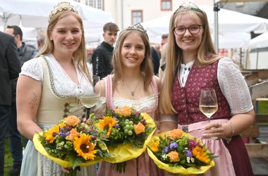 Drei junge Frauen mit langen blonden Haaren im Dirndl und jeweils einem Blumenstrauß mit Sonnenblumen und Trauben in der Hand.