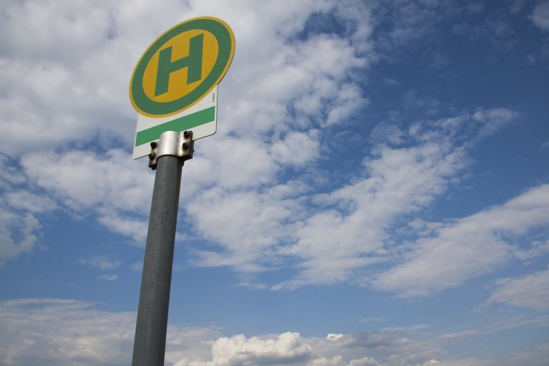 Vor einem blauem Himmel mit weißen Wolken steht ein rundes Bushaltestellenschild mit einem rundem grünen Rahmen und einem grünem "H" auf gelbem Untergrund über einem weißem rechteck mit einem grünem Streifen. Das Schild ist an einer silbernen Metallstange befestigt. 