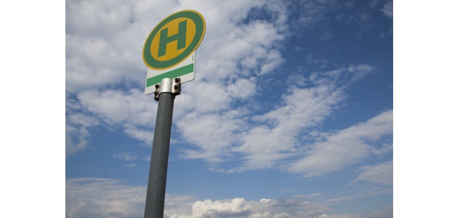 Vor einem blauem Himmel mit weißen Wolken steht ein rundes Bushaltestellenschild mit einem rundem grünen Rahmen und einem grünem "H" auf gelbem Untergrund über einem weißem rechteck mit einem grünem Streifen. Das Schild ist an einer silbernen Metallstange befestigt. 