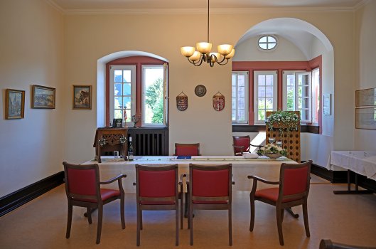 Festlich dekorierter Trausaal mit einer langen Tafel mit beigen Tischdecken, einem aus Holz geschnitztem Rednerpult, mit Blumendeko und Holzstühlen mit rotem Polster. Ein Stuhl steht hinter dem Tisch und vier davor. Im Erker des Raumes steht ein weiterer Stuhl und ein Champagner Rüttelbrett aus Holz. Der Kronleuchter über dem Tisch ist an. Die weißen Sprossenfenster sind zum Teil geöffnet und manche geschlossen. An der Wand hängen alte Gemälde mit goldenen Rahmen.    