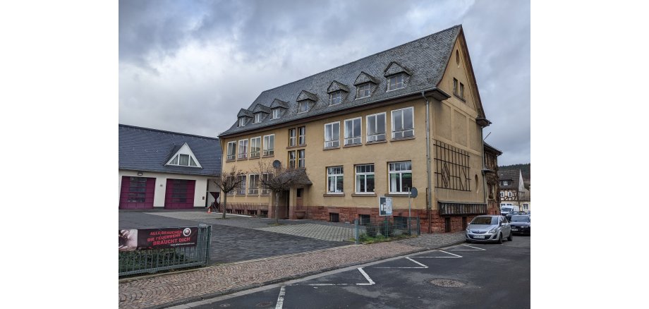 Beiges Gebäude mit Fenstern und Dachfenstern in dem mit schwarzen Ziegeln bedeckten Dach, In dem Gebäude befand sich früher die Schule von Kiedrich. Ebenso sieht man den Hof mit Bäumen und ein Teil der Straße mit einem parkendem Auto. 