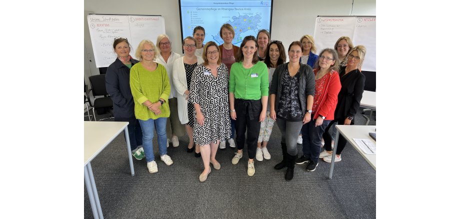 Vor einer Leinwand mit einer Karte, neben der links und rechts mehrere Flip-Charts mit beschrifteten Blättern hängen, stehen  15 Frauen.