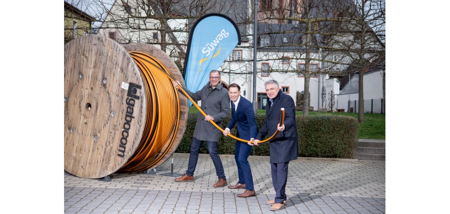 Herr Bürgermeister Steinmacher und zwei Mitarbeiter der Süwag Energie AG, gekleidet mit dunklen Anzugshosen und Mänteln oder Jackets stehen vor dem blauen SÜWAG Dropflag und ziehen an dem Ende eines orangenem Glasfaserkabels, das auf einer großen Holzspule aufgewickelt ist.  