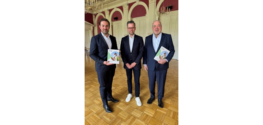 In einem Saal mit Paketboden, Kronleuchtern, weißen Türen und Stuck und weinroter Wandfarbe stehen drei Männer im dunklem Anzug und weißem Hemd mit dem Programmheft des Rheingau Musik Festival in der Hand.