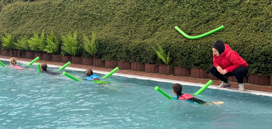 In einem Schwimmbecken in einem Freibad sind 4 Personen im Wasser auf jeweils einer Schwimmnudel. Am Beckenrand erklärt eine Frau mit schwarzer Hose, roter Jacke und schwarzem Kopftuch etwas. Im Hintergrund ist eine grüne Hecke auf der eine weitere Schwimmnudel liegt. 