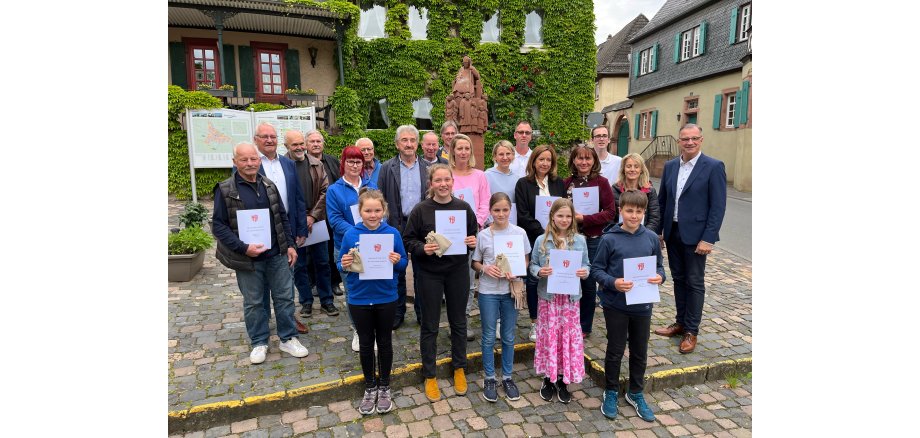 Auf dem Marktplatz mit Pflastersteinen steht eine Gruppe mit Jungen, Mädchen, Frauen und Herren mit Urkunden in der Hand. Herr Bürgermeister Steinmacher steht bei der Gruppe, welche vor einer Werbetafel und einem mit Wein zugewachsenem Haus steht.