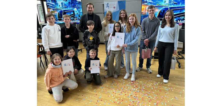 Landrat Sandro Zehner steht mit Schülern, Gewinner und Moderatoren vor dem Banner des Rheingau-Taunus-Kreises und zwei Bildschirmen mit futuristischen Bildern mit Hochhäusern, einer Bahn, Flugelementen und Bäumen. Die Kinder haben zum Teil weiße Urkundenmappen und Geschenktüten in den Händen. Vor der Gruppe liegt Konfetti auf dem Holz-Boden.