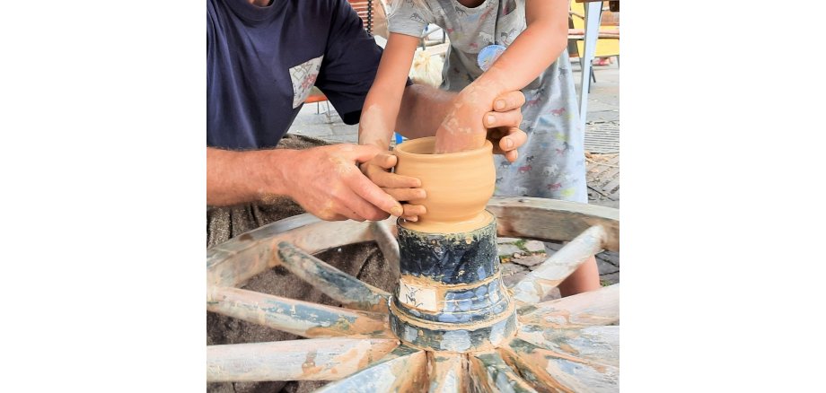 Auf einem Wagenrad aus Holz ist eine Töpfertopf. Zwei Männerhände führen zwei Kinderhände um den Topf zu formen.