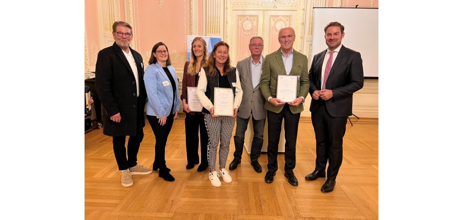 Vier Herren und drei Damen stehen nebeneinander. Drei davon halten eine Urkunde in der Hand. Im Hintergrund eine rosa Wand und Tür, die mit weißem Stuck und goldenen Verzierungen ausgestattet ist.