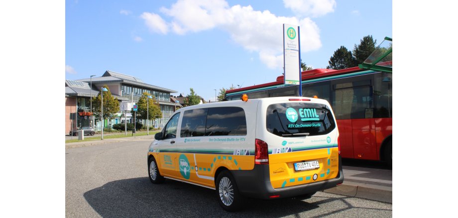 Ein kleiner Bus in weiß und gelb und der türkisen Beschriftung "EMIL", steht an einer Bushaltestelle. Im HIntergrund ein roter Linienbus und ein großes Gebäude mit vielen Fenstern.