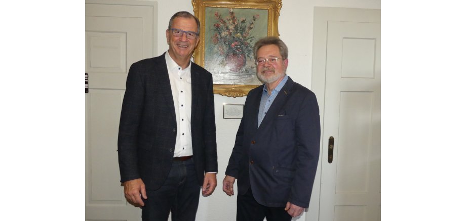 Alfred Zeller mit Bürgermeister Steinmacher, beide im schwarzem Anzug, stehen zwischen zwei weißen Türen und einem Ölgemälde mit einem Blumenstrauss in einer Vase und einem goldenem Bilderrahmen.  