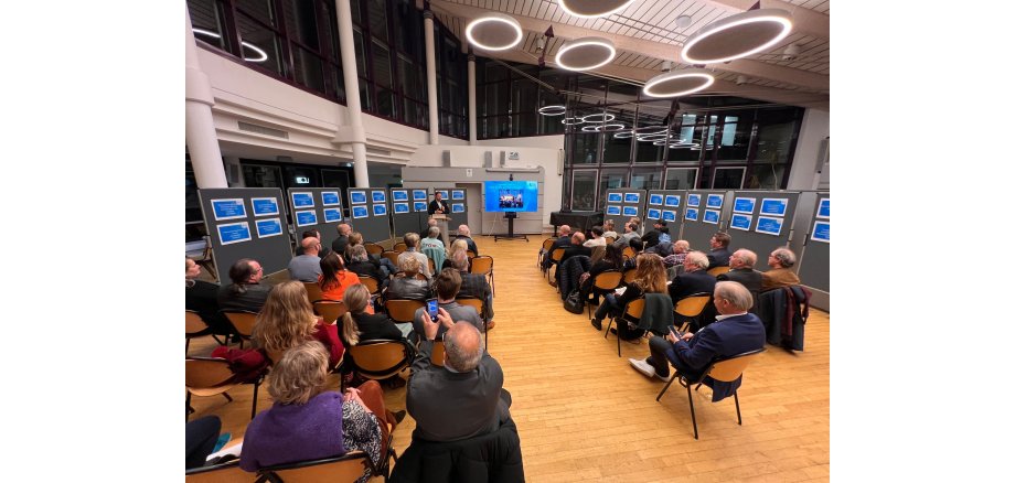 In einem großen, modernen Saal mit einer großen Fensterfront sind viele Stehwände mit Bildern und einem Bildschirm plaziert. In der Mitte sitzen mehrere Personen auf Stühlen und hören dem Redner zu.