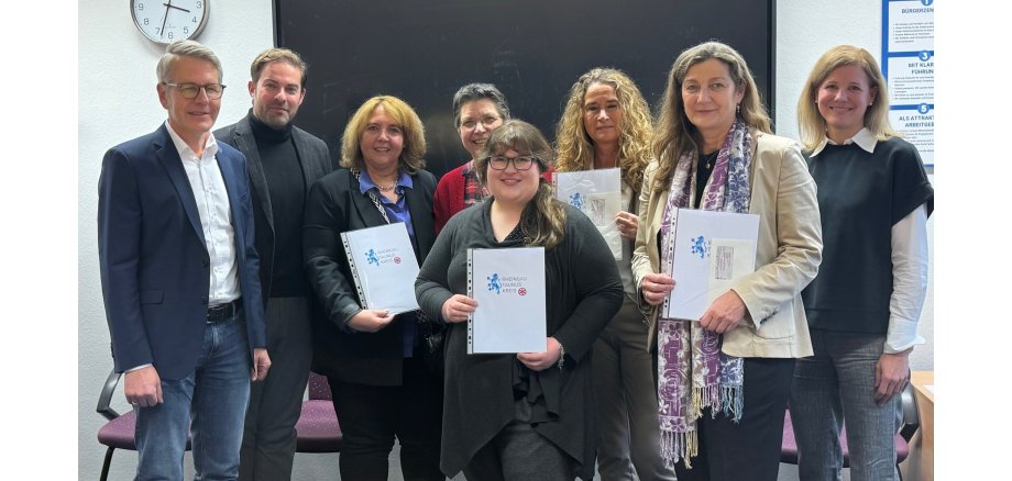 Vor einer Tafel und einer Wanduhr stehen Landrat Zehner mit einem Mann und sechs Frauen, die teilweise Urkunden des Rheingau-Taunus-Kreises in der Hand haben.