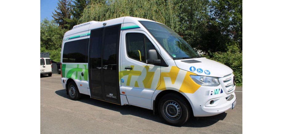 Ein weißer kleiner Bus mit dunklen Scheiben und unterhalb der Scheiben sind in weiß die Buchstaben RTV auf grün-gelben Untergrund. Ebenso ist auf der Motorhaube ein Männchen mit Stock, ein Rollstuhl und ein Kinderwagen in weiß auf blauen Kreisen abgebildet.