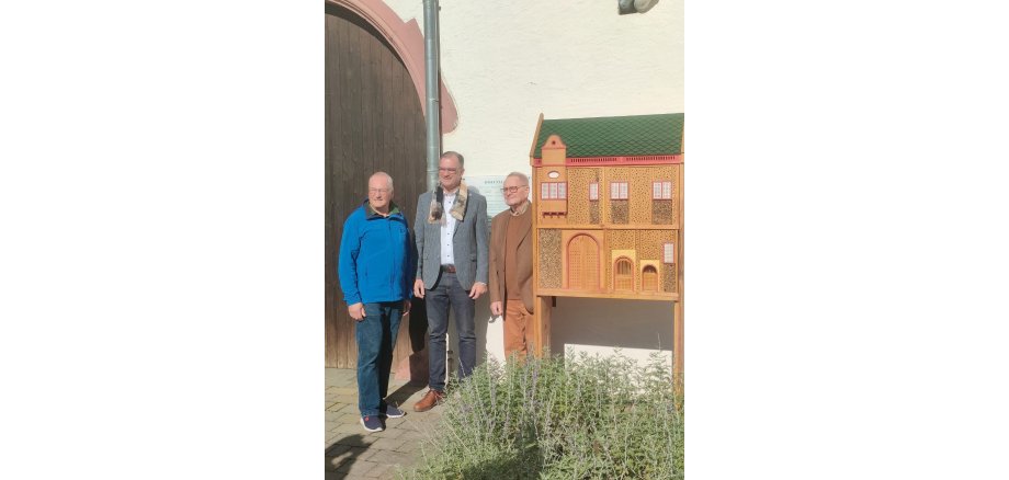 Erster Beigeordneter Rüdiger Wolf (links), Bürgermeister Winfried Steinmacher (Mitte), Dieter Steven (rechts) stehen neben dem neu aufgestelten Insektenhotel, welches mit Holz gebaut wurde und grüne Dachplatten und rote Fenster- und Türrahmen und verschiedenen Erkern besitzt und somit dem Kiedricher Rathaus ähnelt.