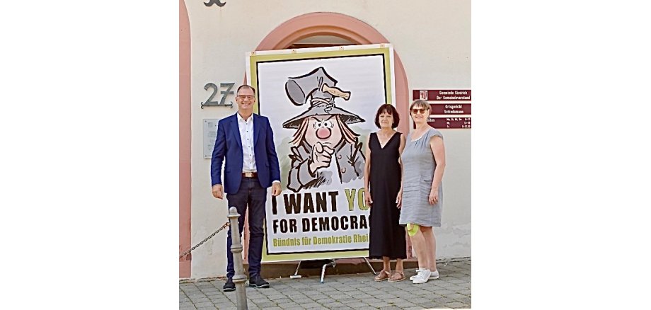 Bürgermeister Steinmacher und zwei Frauen in Kleidern stehen vor dem Rathaus und vor einem Plakat, auf dem ein gezeichneter Mann mit grauem Schlappgut und einem Korkenzieher an der Hutkrempe und einer grauen Jacke abgemildet ist. Er zeigt mit dem Finger auf den Betrachter, schaut grimmig und unter ihm steht "I WANT YOU FOR DEMOCRATY".  