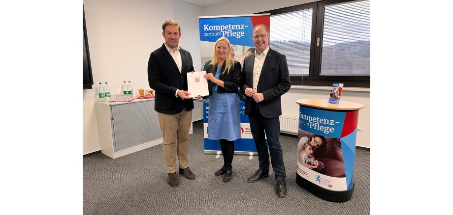 Landrat Sandro Zehner steht neben Staatsministerin Diana Stolz, beide halten zusammen eine Urkundenmappe in der Hand. Rechts davon steht MdL Ingo Schon. Im Hintergrund ist ein Werbebanner und ein Stehtisch mit der Überschrift "Kompetenzzentrum Pflege". 