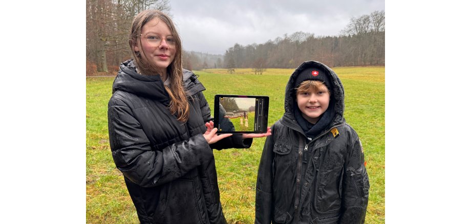 Ein Junge und ein Mädchen stehen mit schwarzen Winterjacken vor einer Wiese, im Hintergrund sind Bäume und halten ein Tablett, auf dem gezeichnet ein römisches Gebäude und zwei Kinder im Römer-Gewand zu sehen ist.