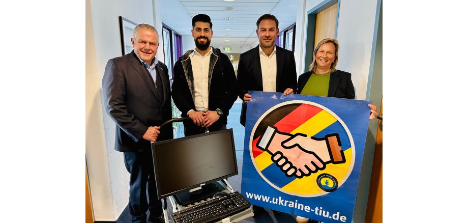 Zwei Herren stehen hinter einem Transportwagen mit einem PC-Bildschirm und einer Tastatur und daneben steht eine Dame und ein Herr, die ein blaues Plakat halten, auf dem in einem Kreis die Farben der deutschen und ukrainischen Fahne abgebildet sind. Zwei Hände machen einen Handschlag und es ist der Hinweis auf www.ukraine-tiu.de.