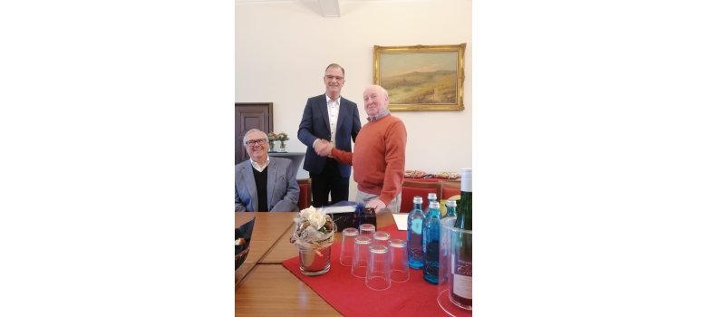 An einem Tisch sitzt Walter Flick, mit weißem Hemd, schwarzem Pullover und grauem Sakko. Auf dem Tisch ist eine kleine rote Tischdecke, auf der ein Blumengesteck und Wassergläser, blaue Wasserflaschen aus Glas und eine Flasche Wein stehen. Daneben steht Bürgermeister Winfried Steinmacher in blauem Anzug und weißem Hemd und gibt Herrn Wilfried Ringel, im orangenem Pullover die Hand.