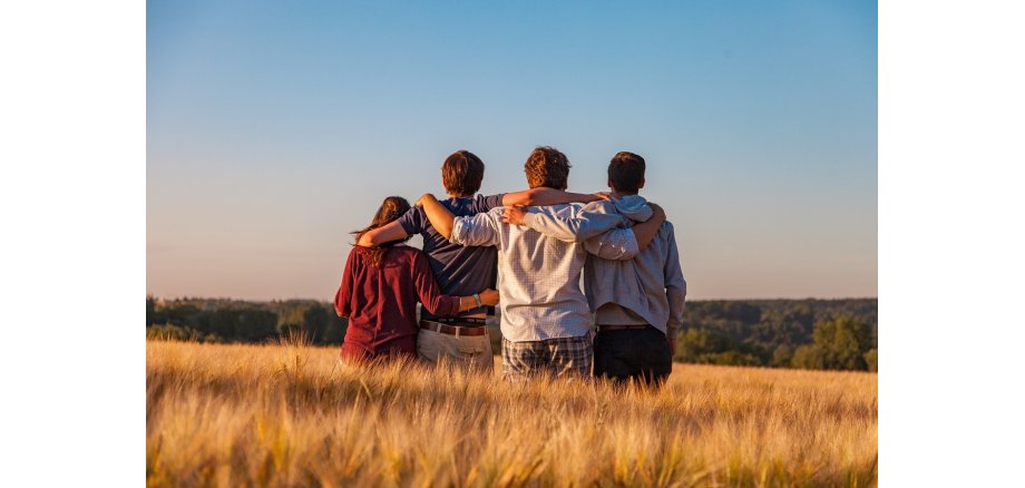 Rückenansicht von vier sich in einer Reihe umarmenden Jugendliche, die auf ein Feld mit Getreide und Bäumen blicken.