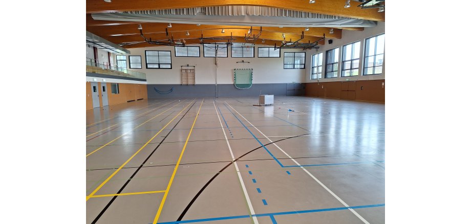 Sporthalle mit diversen farbigen Linien für Sportfelder auf dem Boden, mit einer an der Wand hängenden Sprossenwand und einem zusammengeklappten, ebenso an der Wand hängendem Fussballtor. Es sind mehrere Fenster zu sehen.