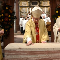 Um den Altar steht ein Pfarrer, ein Diakon und ein Bischof in Gewändern. Im Hintergrund ist verschwommen der Chor der Kiedricher Chorbuben in den Chorröcken zu erkennen.