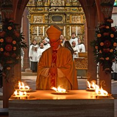 Ein Bischof steht am Altar, auf dem fünf Feuerschalen mit brennendem Feuer stehen. Im HIntergrund ist der Chor der Kiedricher Chorbuben, die Männer und die Kinder im weißen Chorrock sowie der Frauenchor in schwarz gekleidet.