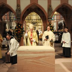 Um den Altar steht ein Pfarrer, ein Diakon und ein Bischof in Gewändern. Der Bischof weiht den Altar. Im Hintergrund ist der Chor der Kiedricher Chorbuben in den Chorröcken zu erkennen.
