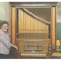 Eine ältere frau, Wilma Scholl, mit kurzen braun gelockten Haaren und einer Brille steht an einer Orgel mit Orgelpfeifen aus Holz.