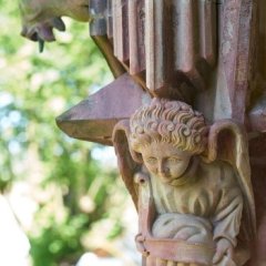 Eine Engelsfigur aus Sandstein an der Kanel der Kiedricher Michaelskapelle mit einem Band in den Händen. Im Hintergrund ist das Profil eines Ziegenbocks mit en Hörnen und dem Ziegenbart zu erkennen. Verschwobben sind dahinter die Blätter der Bäume dargestellt.